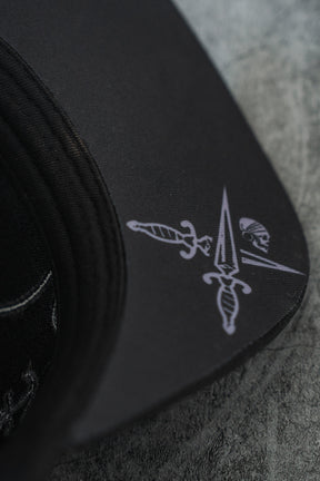 Close-up of the Never Surrender Limited Edition Snapback by Relentless Betrayal, featuring a printed design with two crossed daggers, a pointed spear, and a small skull on the brim, displayed on a grey textured surface.