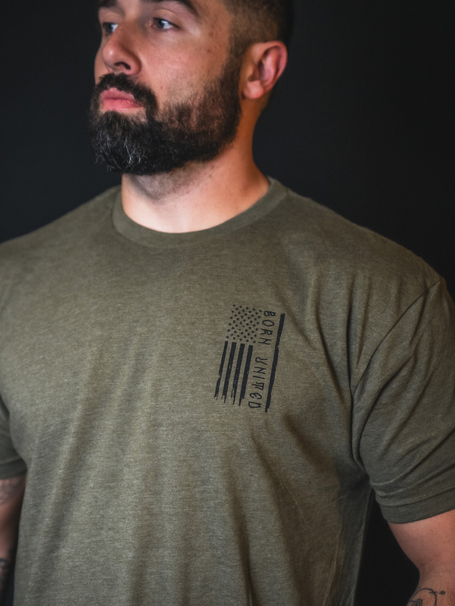 A bearded man wears the limited-edition Tactical Grinch Tee by Born United Private Label—an olive green shirt featuring an American flag and the phrase “Born Humble” on the left chest—standing against a dark background and exemplifying true patriot fashion.