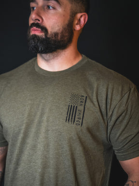 A bearded man wears the limited-edition Tactical Grinch Tee by Born United Private Label—an olive green shirt featuring an American flag and the phrase “Born Humble” on the left chest—standing against a dark background and exemplifying true patriot fashion.