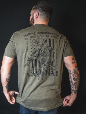 A man with tattoos wears the olive green Tactical Grinch Tee by Born United Private Label, a limited-edition shirt featuring a soldier graphic and the phrase, “Making Christmases so yours can be merry. Born Untamed.” This piece combines festive spirit with bold patriotic style.