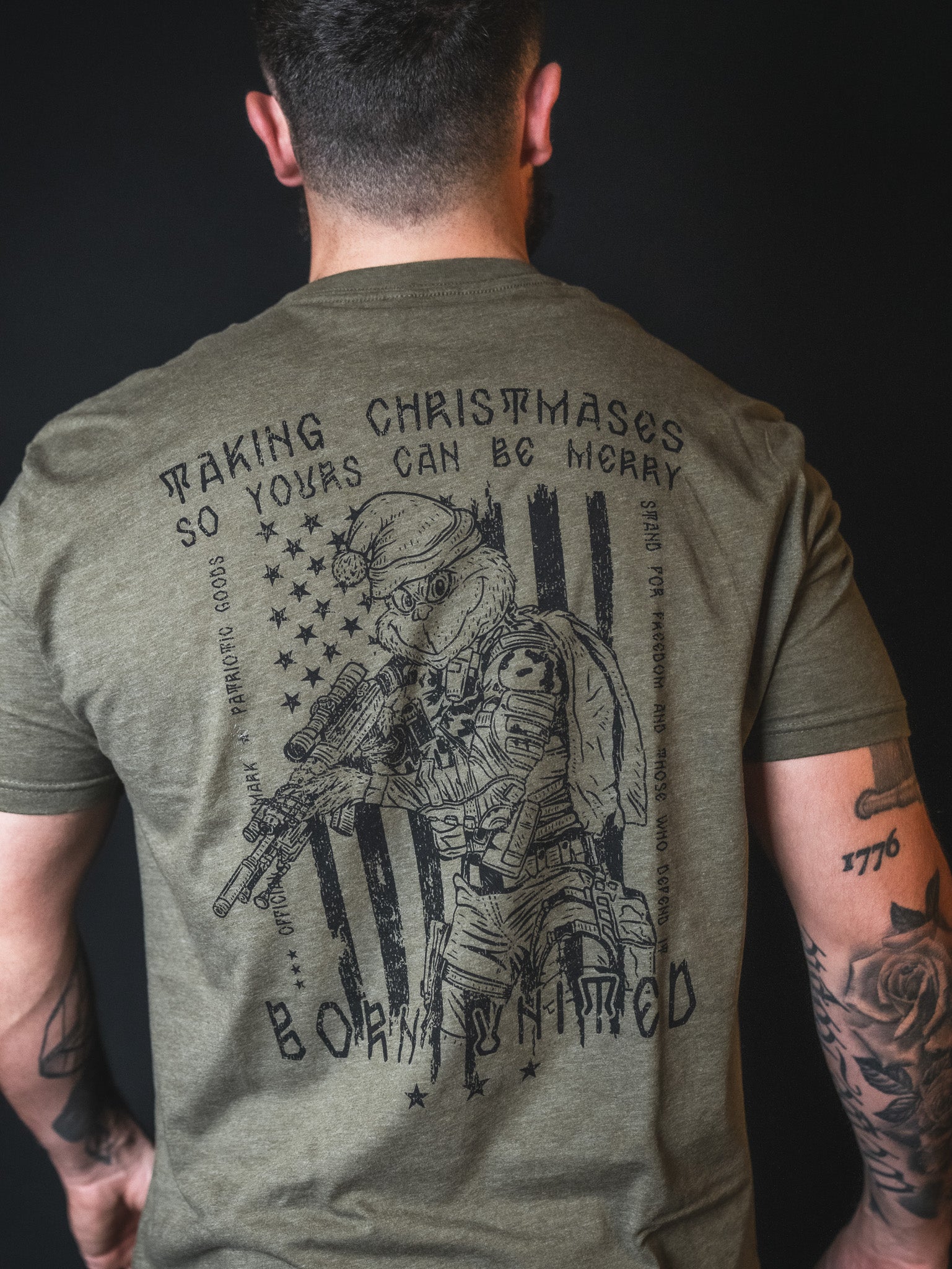 A man wearing the Tactical Grinch Tee by Born United Private Label—a limited-edition olive green shirt featuring a graphic of an armed soldier, an American flag, and the text “Making Christmases so yours can be merry. Born Untamed.” This piece fuses patriotic style with Christmas spirit for a truly bold statement.