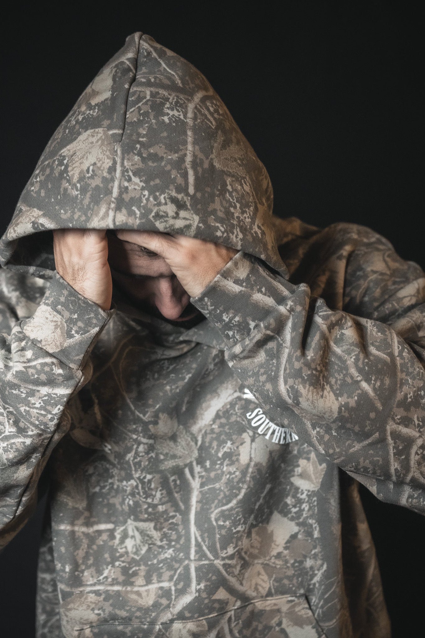 A person in a Mucker Bucket Mallard Deadwood Camo Hoodie with the hood up covers their face with both hands, standing against a dark background.