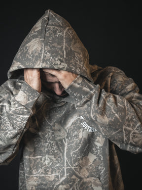 A person in a Mucker Bucket Mallard Deadwood Camo Hoodie with the hood up covers their face with both hands, standing against a dark background.