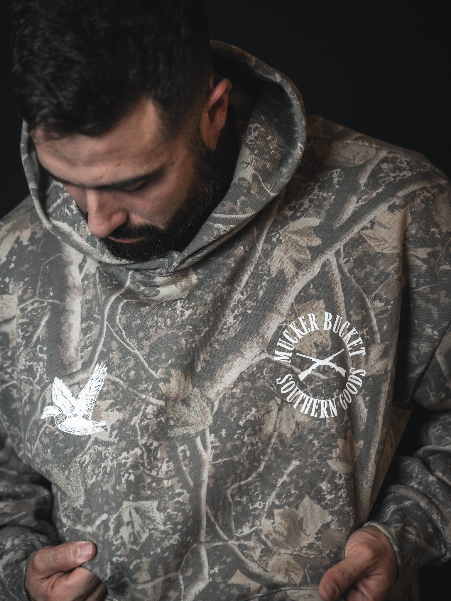 A bearded man looks down, wearing the Mallard Deadwood Camo Hoodie from Mucker Bucket, featuring a flying duck graphic. He holds the sides of this comfortable hoodie for a relaxed fit.