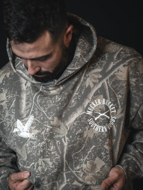 A bearded man looks down, wearing the Mallard Deadwood Camo Hoodie from Mucker Bucket, featuring a flying duck graphic. He holds the sides of this comfortable hoodie for a relaxed fit.
