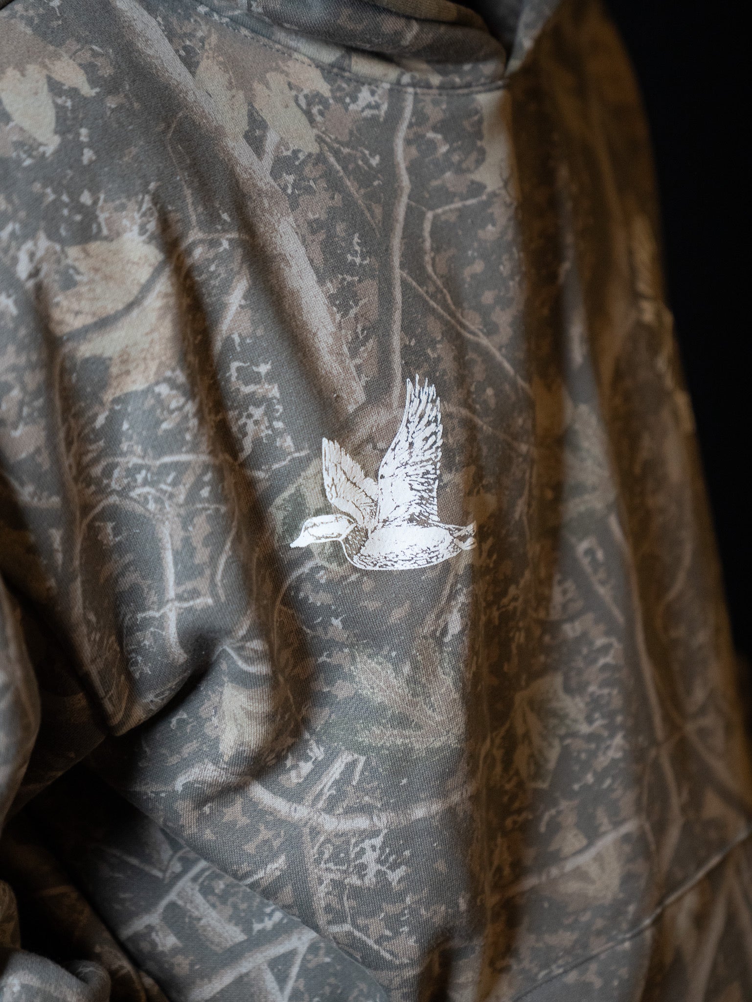 A close-up of the Mucker Bucket Mallard Deadwood Camo Hoodie, showcasing its trendy camouflage pattern and an embroidered white duck in flight on the back.