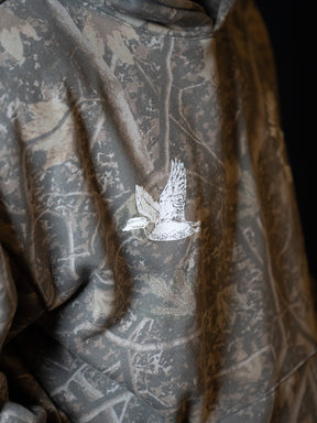 A close-up of the Mucker Bucket Mallard Deadwood Camo Hoodie, showcasing its trendy camouflage pattern and an embroidered white duck in flight on the back.