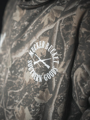 A close-up of the Mucker Bucket Mallard Deadwood Camo Hoodie featuring a white MUCKER BUCKET SOUTHERN GOODS logo and two crossed rifles—perfect to pair with your favorite premium relaxed fit styles.