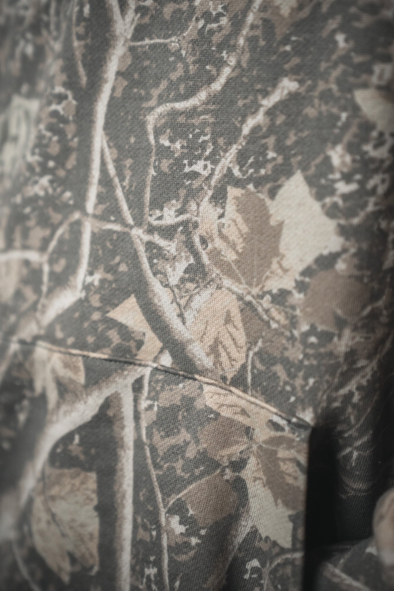 Close-up of the Mucker Bucket Mallard Deadwood Camo Hoodie features brown leaves and tree branches in earthy tones, highlighting its textured fleece fabric and detailed stitching.
