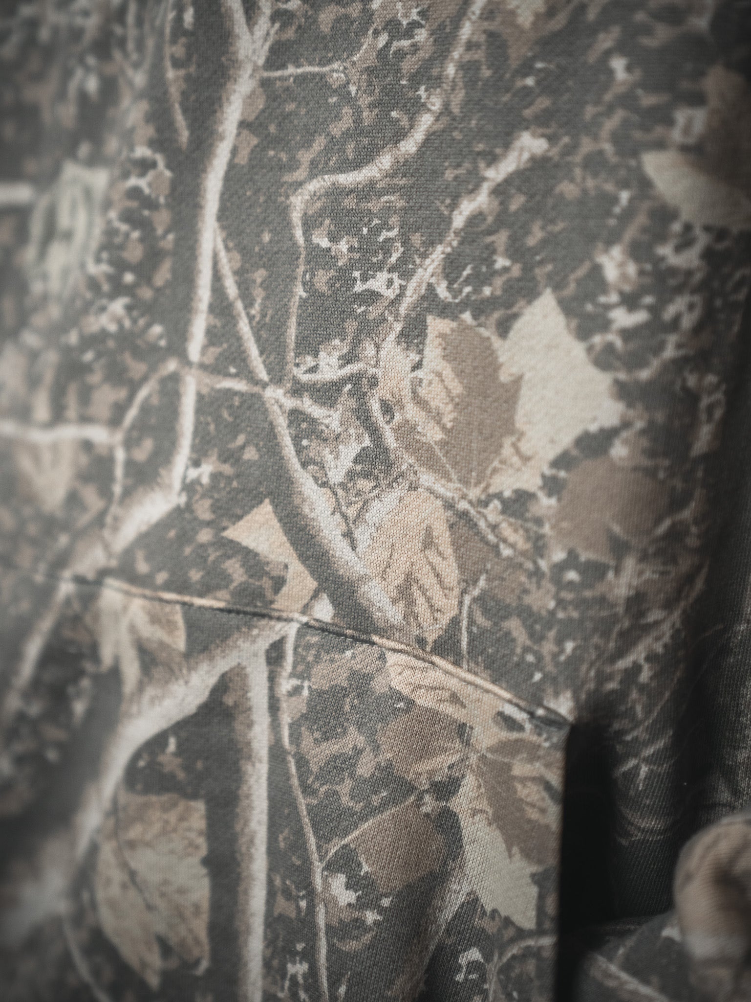 Close-up of the Mucker Bucket Mallard Deadwood Camo Hoodie features brown leaves and tree branches in earthy tones, highlighting its textured fleece fabric and detailed stitching.