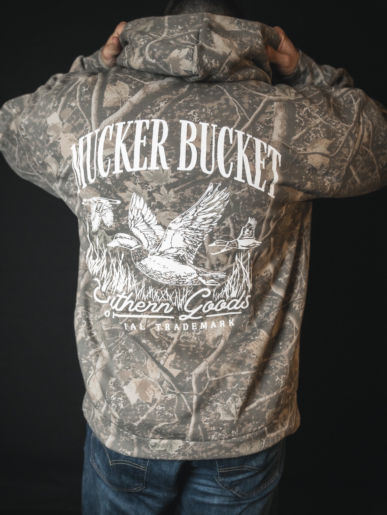A person faces away in a Mallard Deadwood Camo Hoodie by Mucker Bucket, featuring stacking ducks, reeds, and a dog graphic on the back, plus Southern Goods and TRADEMARK.