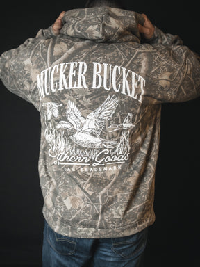 A person faces away in a Mallard Deadwood Camo Hoodie by Mucker Bucket, featuring stacking ducks, reeds, and a dog graphic on the back, plus Southern Goods and TRADEMARK.