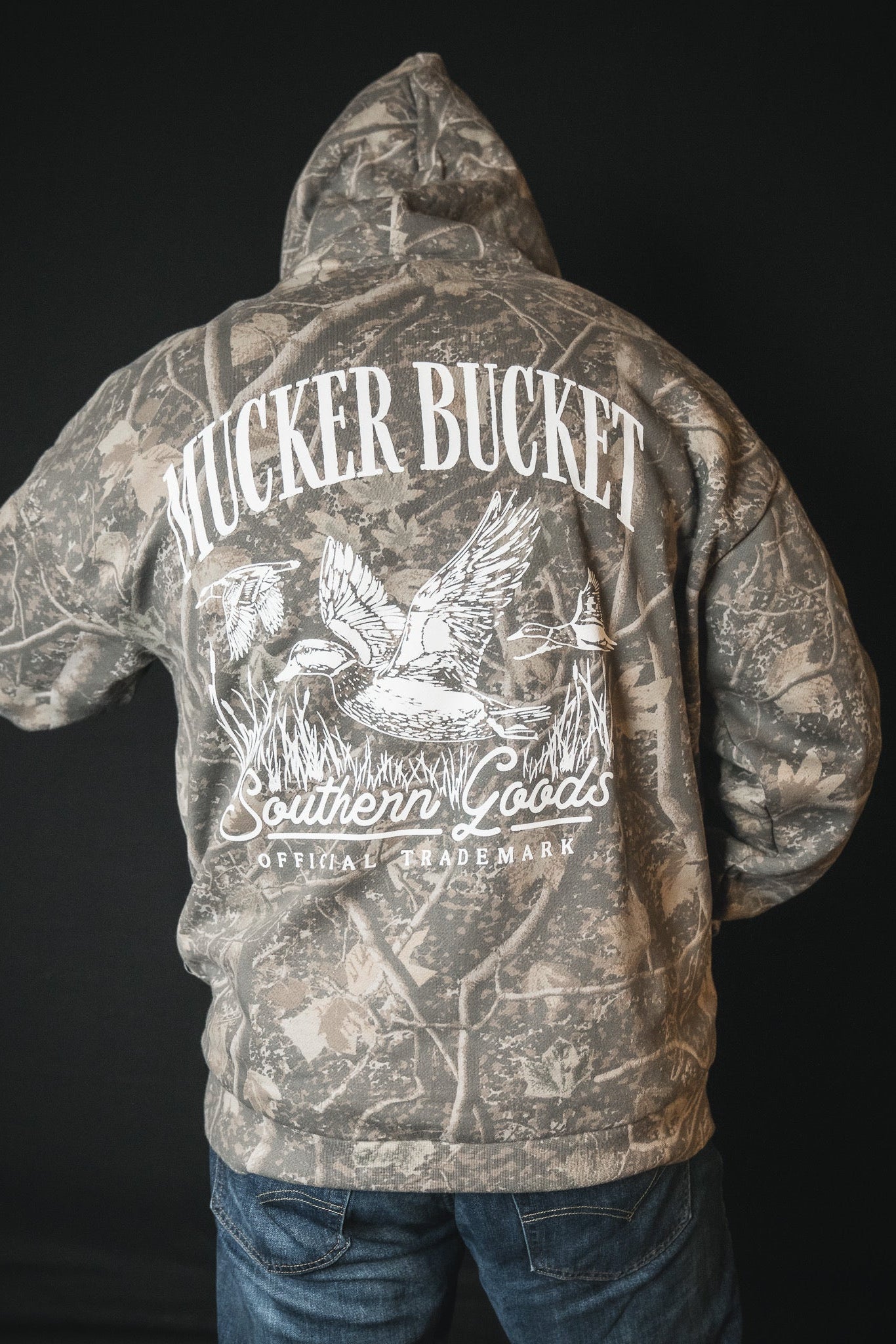 A person wearing the Mucker Bucket Mallard Deadwood Camo Hoodie, featuring a hooded camouflage design and white graphics on the back with "MUCKER BUCKET," "Southern Goods," "Official Trademark," flying ducks, and reeds.