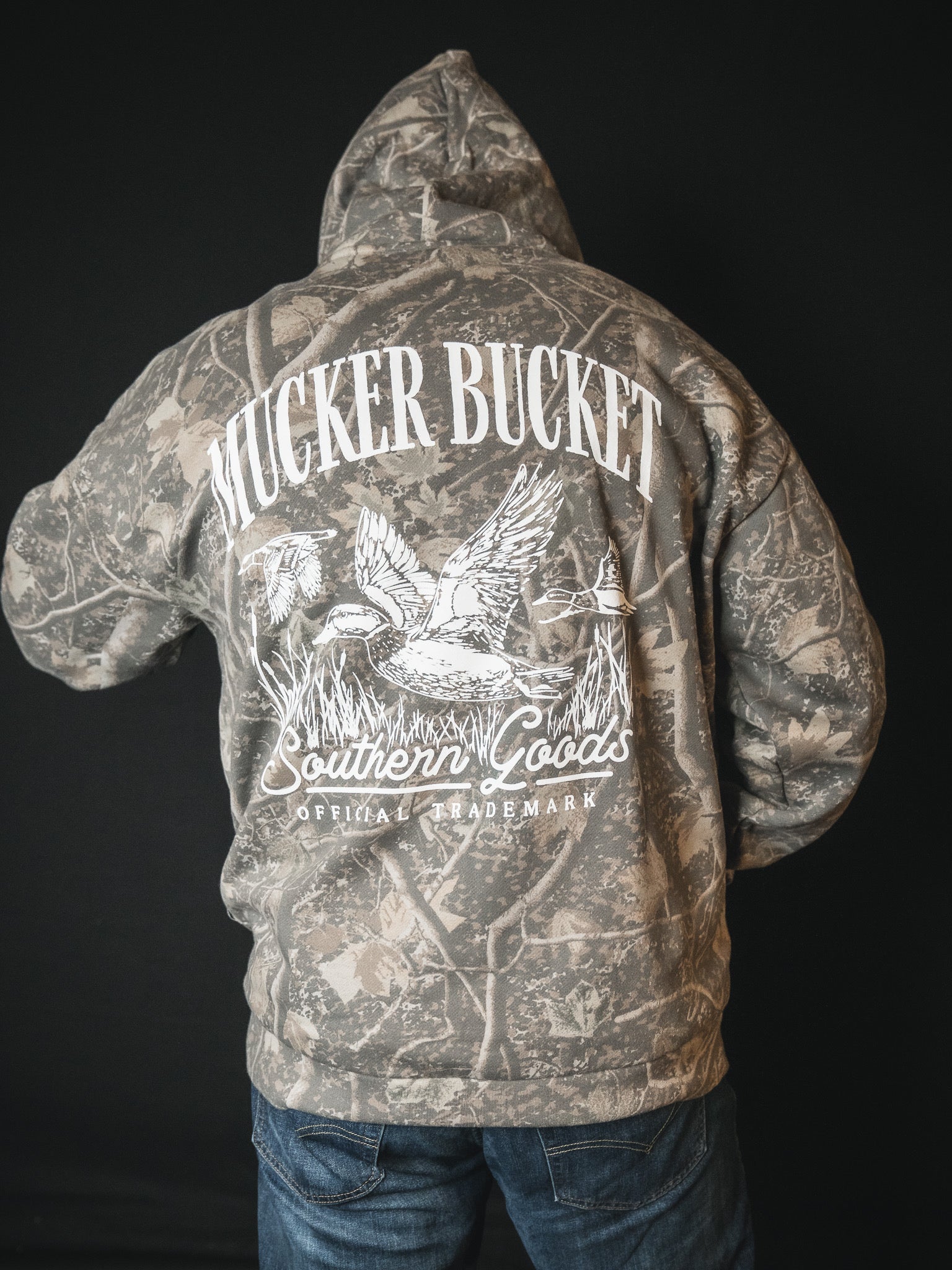 A person wearing the Mucker Bucket Mallard Deadwood Camo Hoodie, featuring a hooded camouflage design and white graphics on the back with "MUCKER BUCKET," "Southern Goods," "Official Trademark," flying ducks, and reeds.
