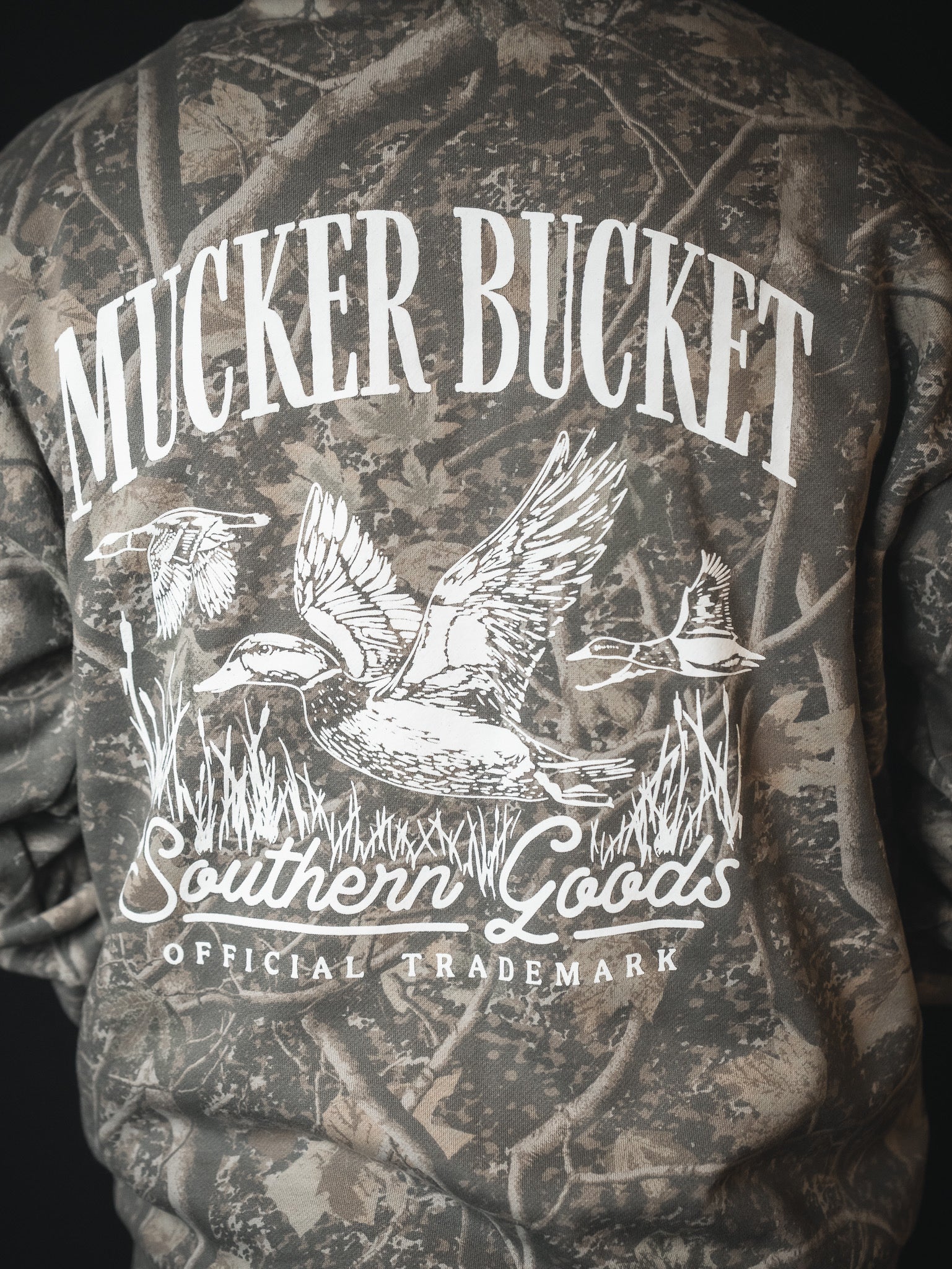 Someone wears the Mallard Deadwood Camo Hoodie by Mucker Bucket, featuring white MUCKER BUCKET text above a graphic of three ducks with grass, and "Southern Goods Official Trademark" printed below the image.