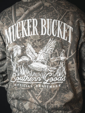 Someone wears the Mallard Deadwood Camo Hoodie by Mucker Bucket, featuring white MUCKER BUCKET text above a graphic of three ducks with grass, and "Southern Goods Official Trademark" printed below the image.
