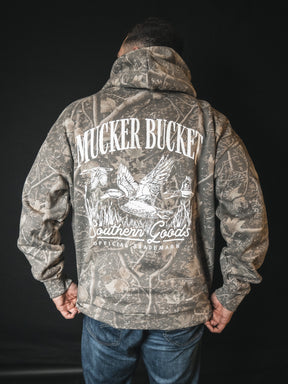 A person wears a Mallard Deadwood Camo Hoodie by Mucker Bucket—featuring relaxed fit and illustrated ducks in flight—standing against a plain black background.