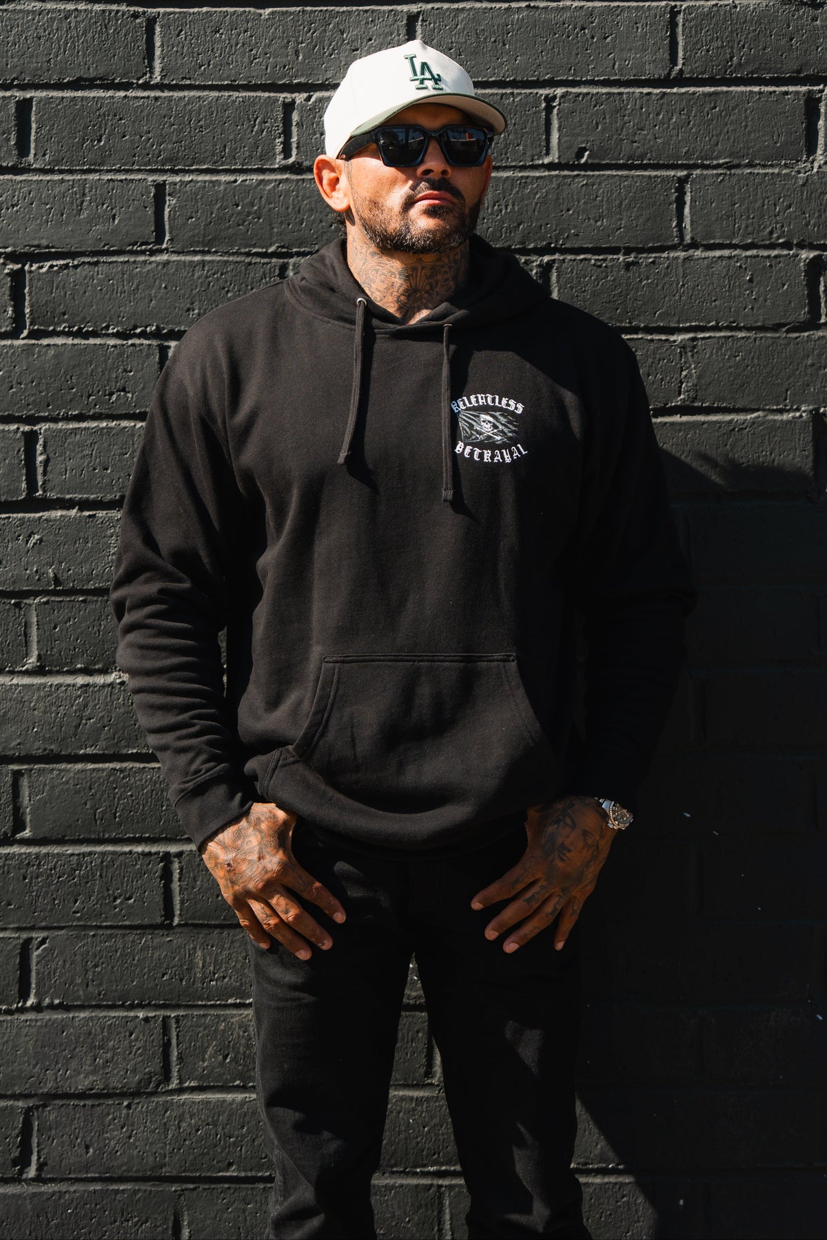 A man in a Raise the Jolly Roger Hoodie by Relentless Betrayal, black pants, and a white cap stands against a black brick wall, facing forward with hands at his sides.