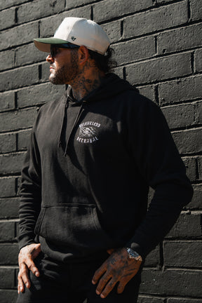 A man wearing the Raise the Jolly Roger Hoodie by Relentless Betrayal and a white cap leans against a black brick wall, capturing bold pirate streetwear vibes.