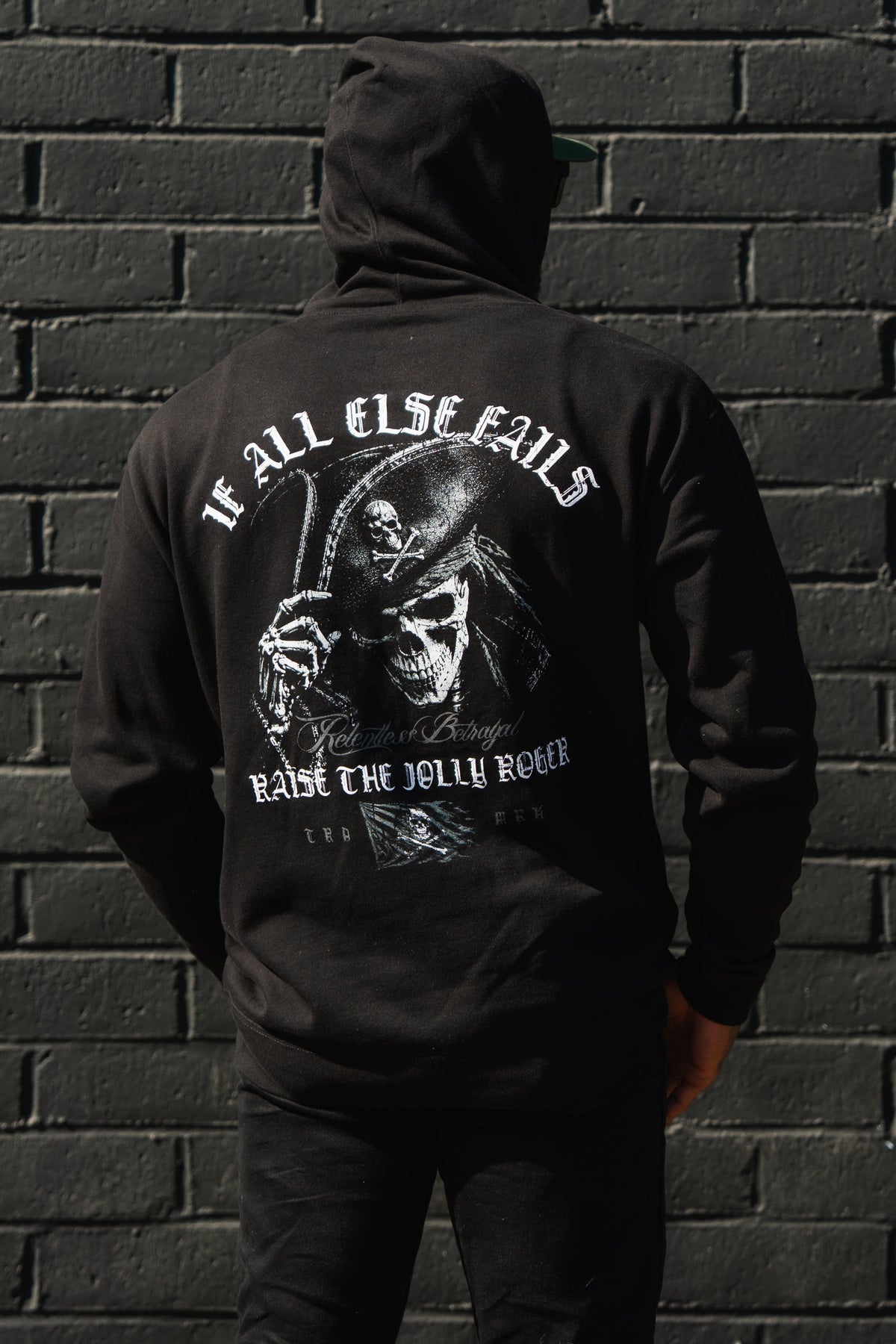 Wearing a Raise the Jolly Roger Hoodie by Relentless Betrayal, someone with pirate streetwear style stands before a black brick wall.