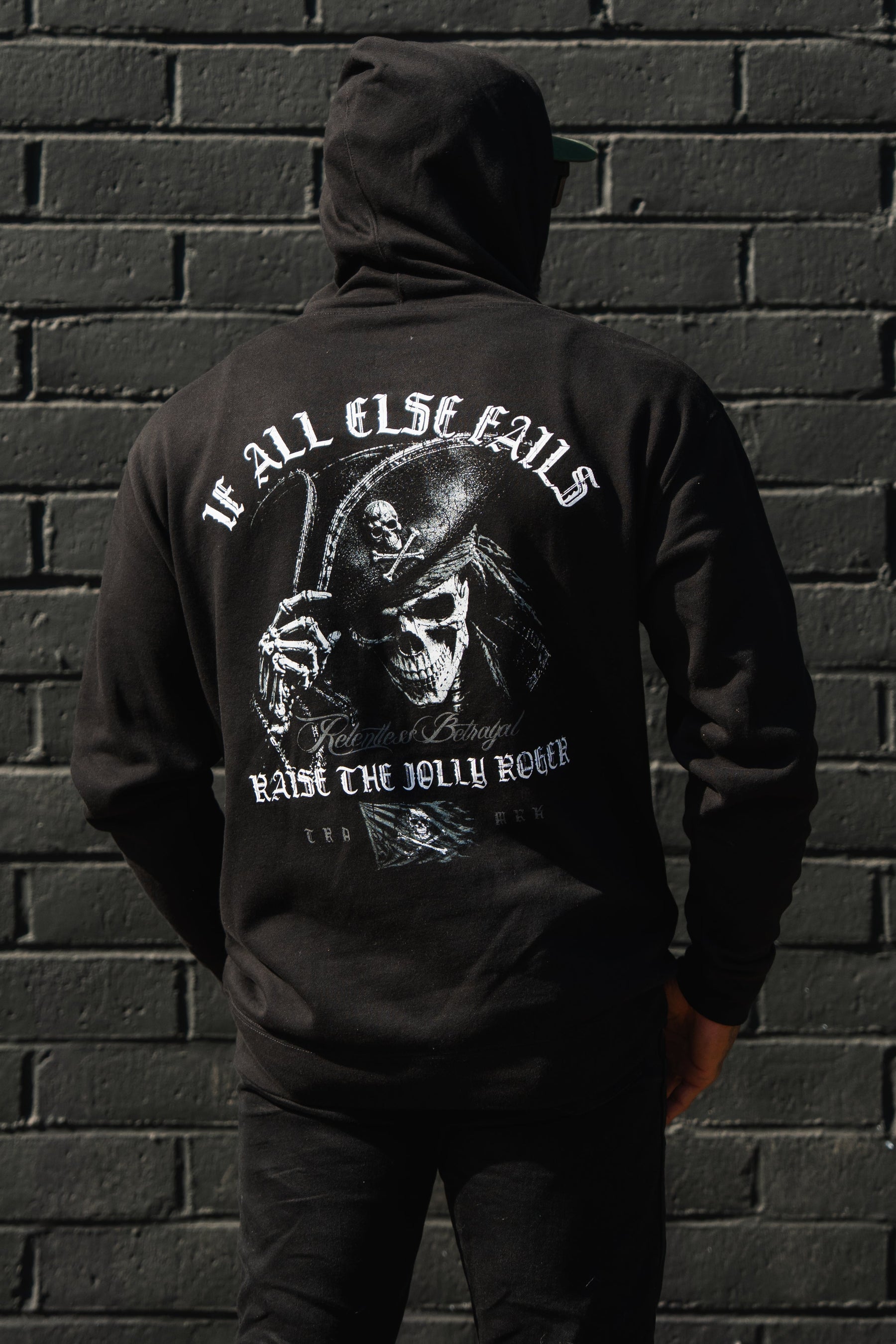Wearing a Raise the Jolly Roger Hoodie by Relentless Betrayal, someone with pirate streetwear style stands before a black brick wall.