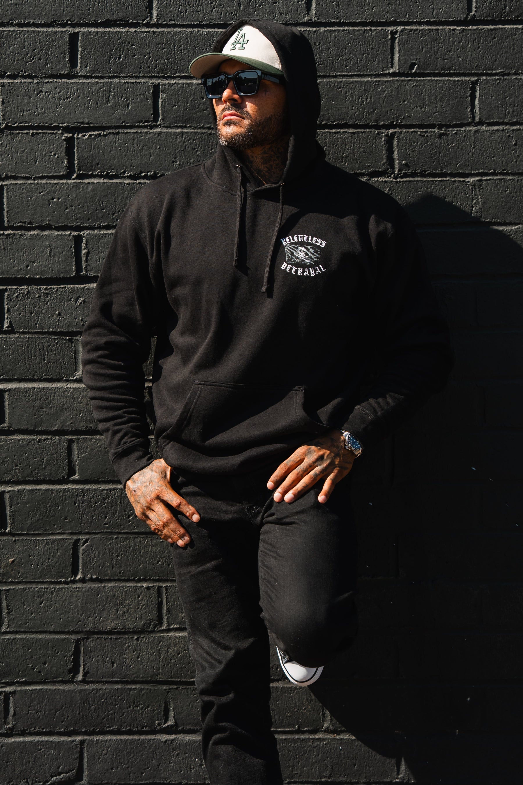 A man in a Raise the Jolly Roger Hoodie by Relentless Betrayal and cap leans against a black brick wall, wearing sunglasses and black jeans, one leg bent.