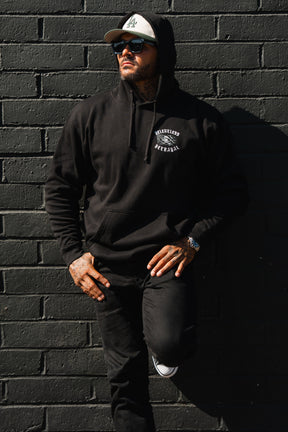 A man in a Raise the Jolly Roger Hoodie by Relentless Betrayal and cap leans against a black brick wall, wearing sunglasses and black jeans, one leg bent.
