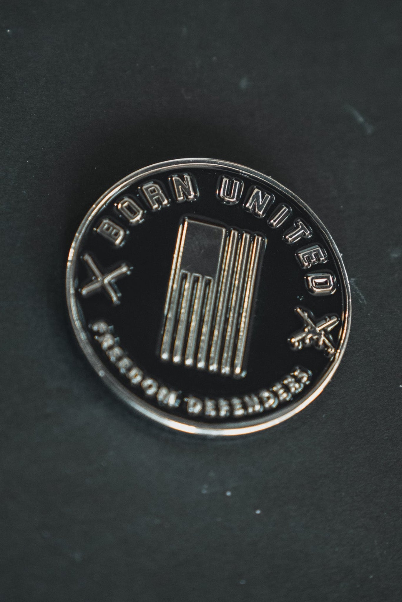 A round, metallic pin on a dark surface features an American flag at the center with Born United and Freedom Defenders text. Two cross motifs symbolize unity for a brighter future. Product: Saints and Sinners Challenge Coin by Born United Private Label.