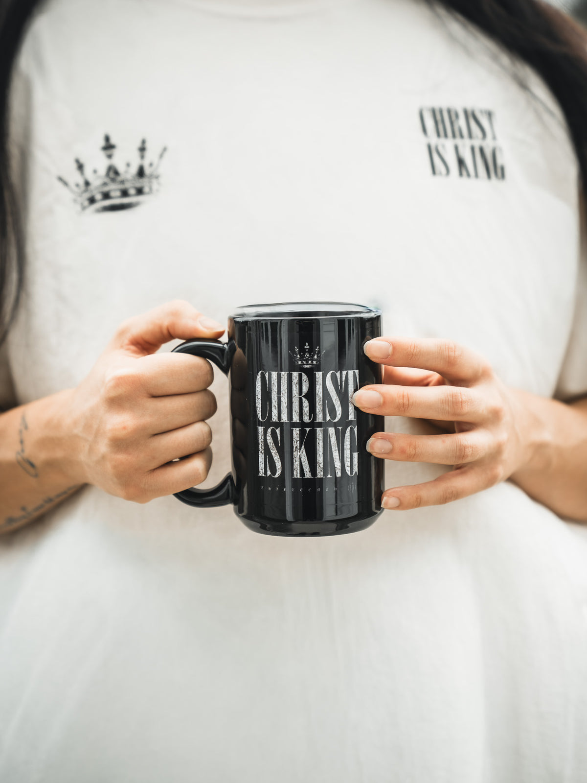 A person holds the 13th Christ is King Mug, a black cup with the Christian phrase and crown graphic, while wearing a matching white "Christ Is King" shirt. Only their hands and torso are seen.