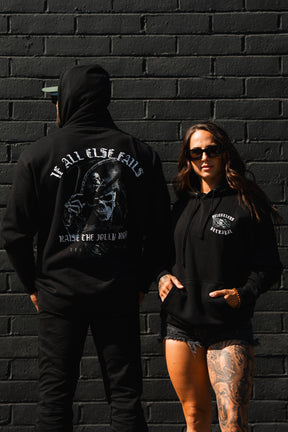 Two people in black Raise the Jolly Roger Hoodies by Relentless Betrayal show off the pirate skull graphic and “If All Else Fails, Raise the Jolly Roger” text.