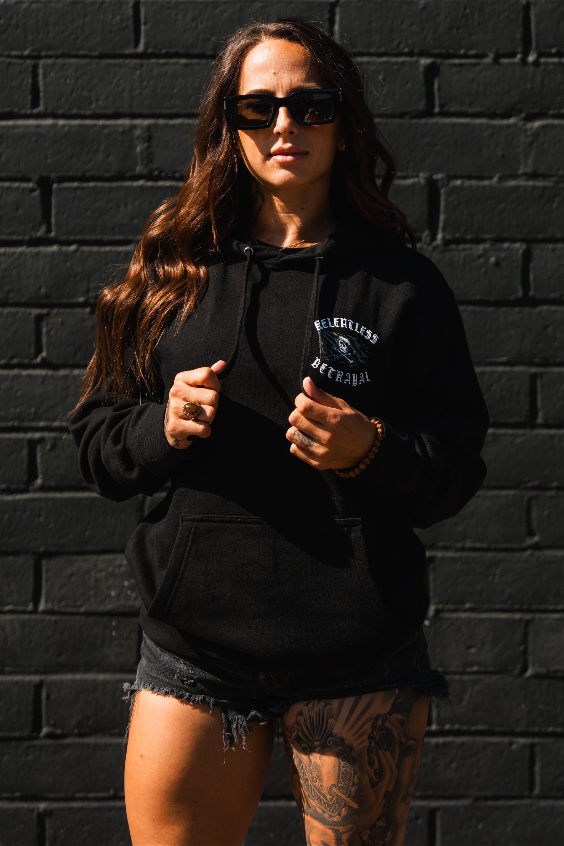 A woman in the Raise the Jolly Roger Hoodie by Relentless Betrayal stands against a black brick wall, holding the strings, with sunglasses and one tattooed leg visible.