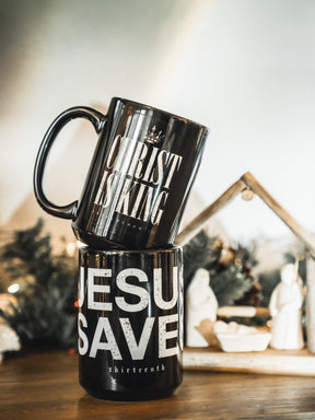 Two black 13th mugs are stacked on a wooden surface—the top reads “CHRIST IS KING,” and the Jesus Saves Mug is beneath. A blurred nativity scene and Christmas greenery add a festive touch in the background.