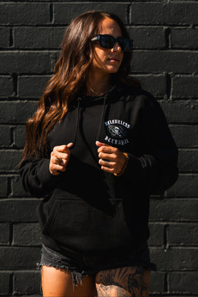 Woman in sunglasses and black shorts stands before a dark brick wall, wearing the Raise the Jolly Roger Hoodie by Relentless Betrayal for bold pirate streetwear vibes.