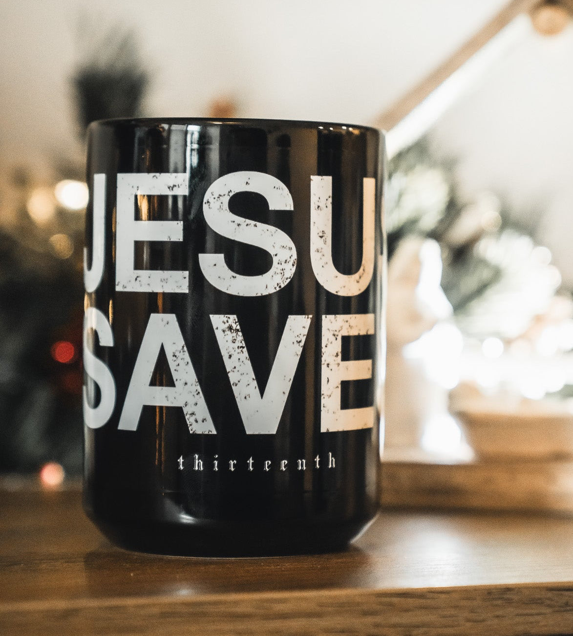 A black 13th Jesus Saves Mug with bold white lettering sits on a wooden surface, with a blurred nativity scene and festive Christmas decorations creating a warm background.