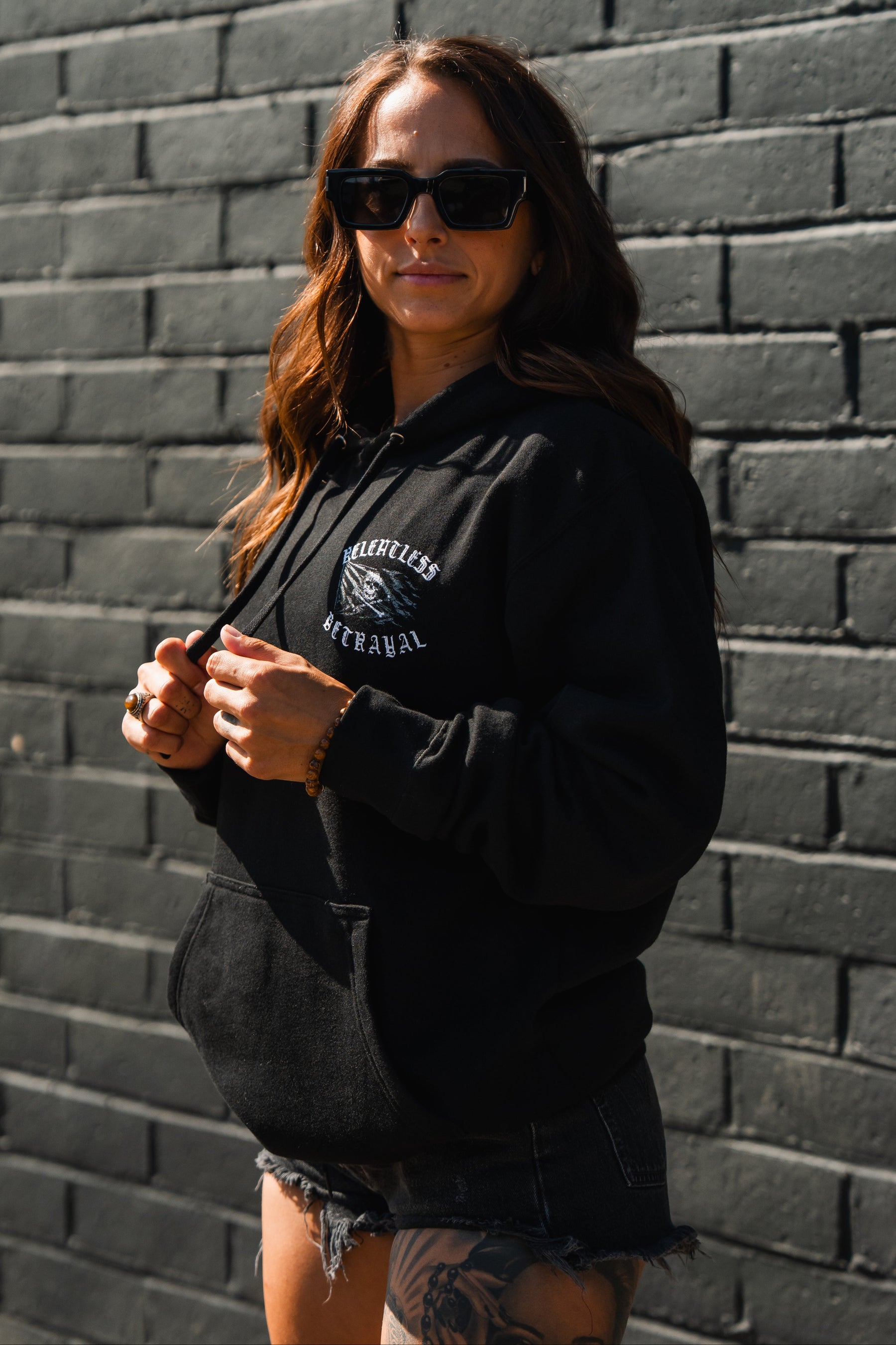 A woman in sunglasses and black Raise the Jolly Roger Hoodie by Relentless Betrayal stands against a brick wall, wearing black shorts and showing a thigh tattoo.