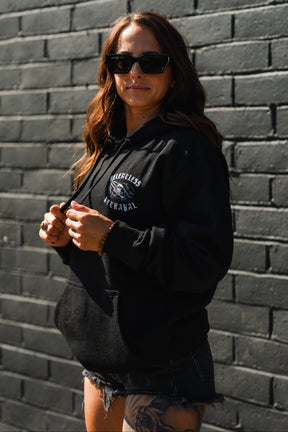 A woman in sunglasses and black Raise the Jolly Roger Hoodie by Relentless Betrayal stands against a brick wall, wearing black shorts and showing a thigh tattoo.