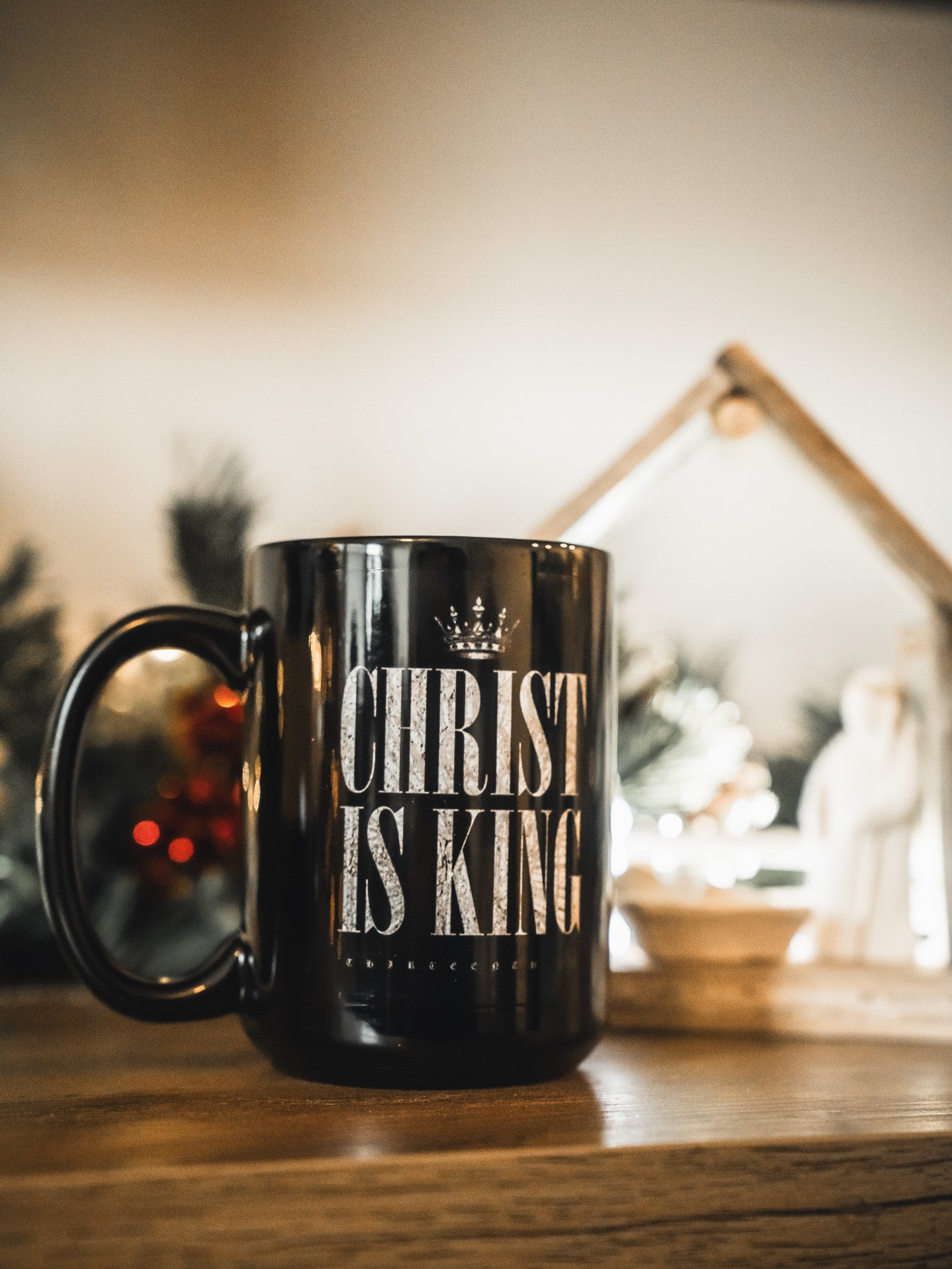 The 13th Christ is King Mug, featuring bold white text on black, rests on a wooden surface with a blurred nativity scene and holiday decor in the background.