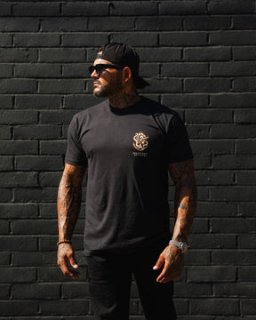 A man in a Relentless Betrayal No Safe Harbor Premium T-Shirt, sunglasses, and a backward cap stands by a black brick wall, looking to the side with relaxed arms.