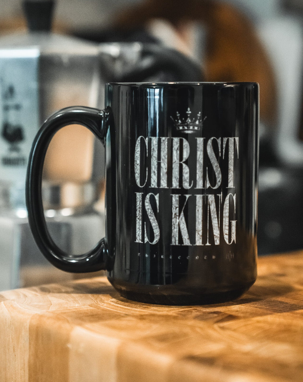 The Christ is King Mug by 13th, featuring bold white CHRIST IS KING lettering on black, sits on a wooden countertop with a blurred silver stovetop espresso maker in the background, expressing Christian faith.