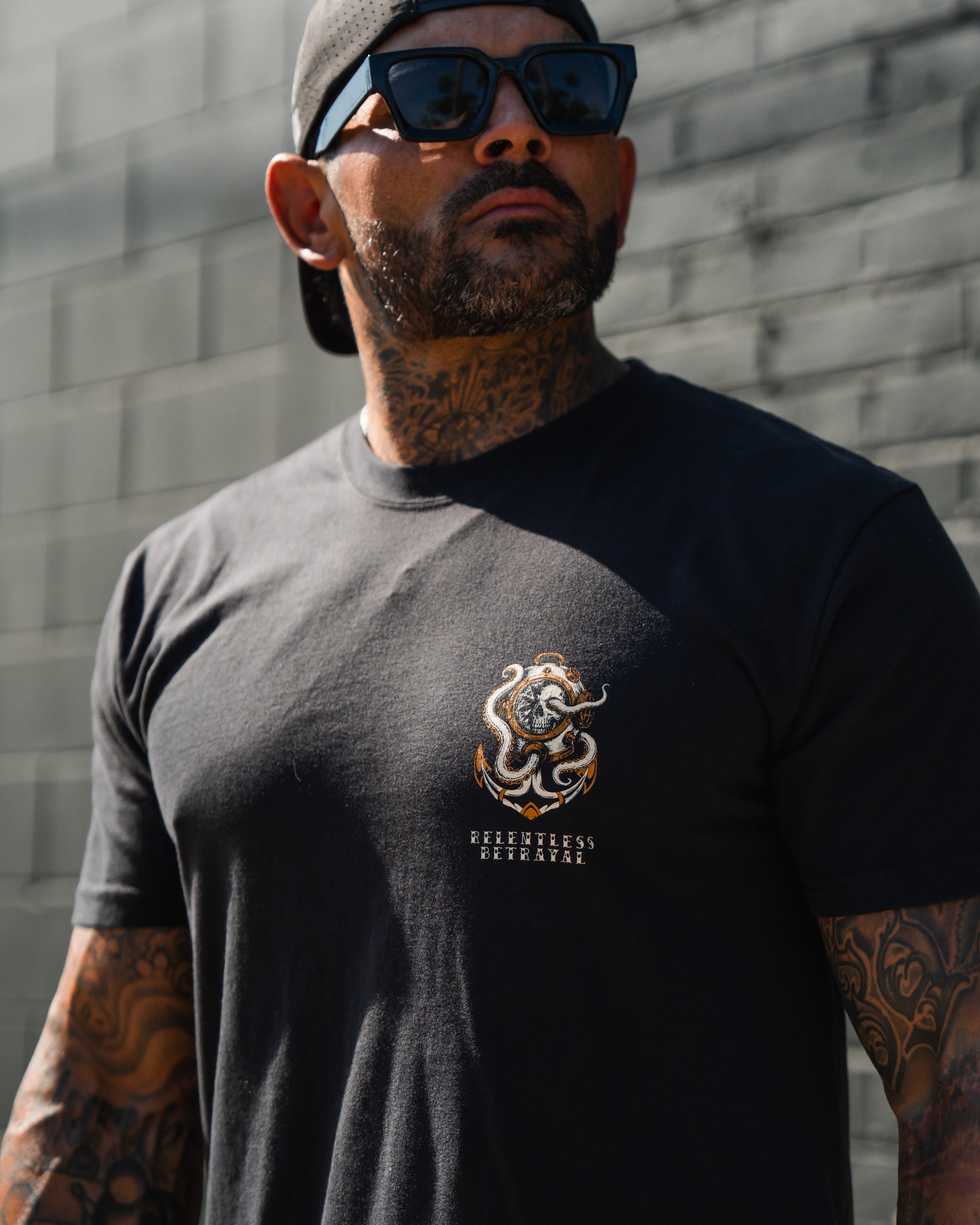Tattooed man in sunglasses, black cap, and No Safe Harbor Premium T-Shirt by Relentless Betrayal stands against a gray brick wall.