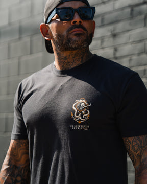 Tattooed man in sunglasses, black cap, and No Safe Harbor Premium T-Shirt by Relentless Betrayal stands against a gray brick wall.