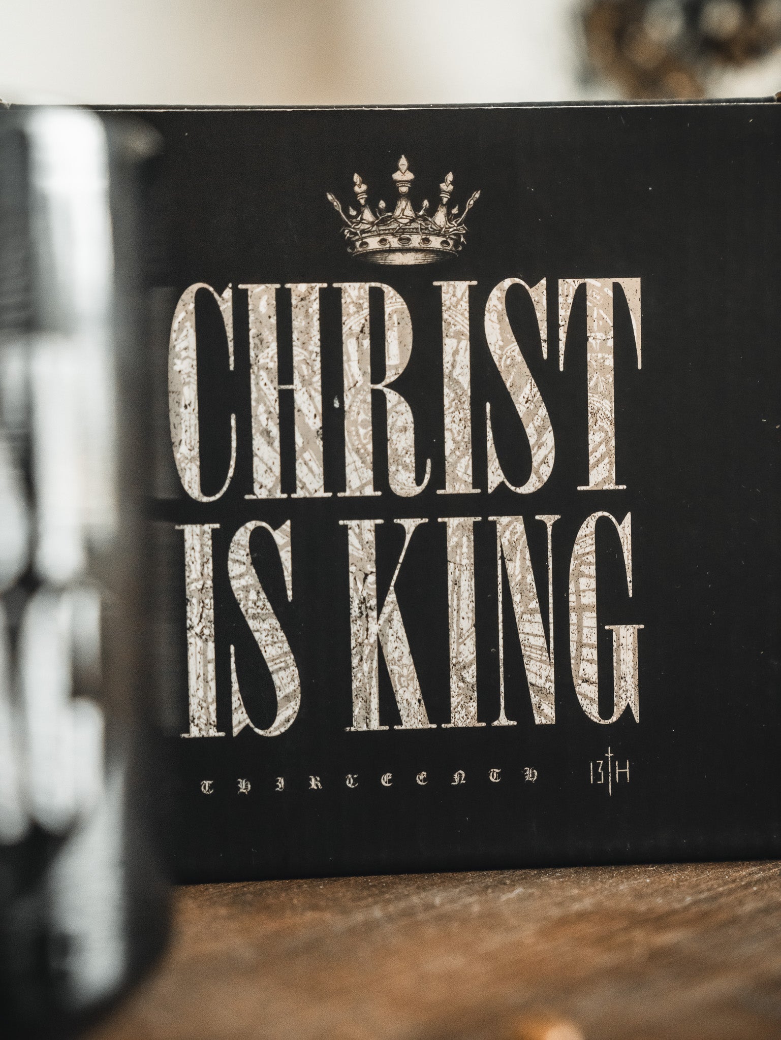 The Christ is King Mug by 13th features bold, distressed silver lettering and a crown graphic on a black background, with “THIRTEENED 13TH” in small text at the bottom. The mug celebrates Christian faith and is shown on a wooden surface.