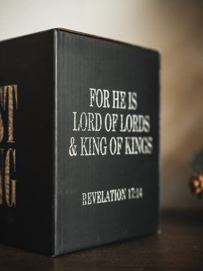 The Christ is King Mug by 13th features a black design with white text—FOR HE IS LORD OF LORDS & KING OF KINGS REVELATION 17:14—displayed on a wooden surface, serving as a daily reminder of Christ’s kingship.