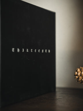 A black Christ is King Mug by 13th stands upright on a dark surface. A pine cone on the right contributes to a solemn mood that echoes the mug’s bold message.