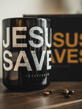 The 13th Jesus Saves Mug in black, featuring bold white text, rests on a wooden surface with coffee beans. A blurred background with yellow accents adds warmth and inspiration to the scene.