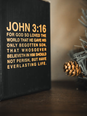 A close-up shows a black box with “John 3:16” in yellow beside a pinecone, an evergreen branch, and the 13th brand’s Jesus Saves Mug on a wooden surface.