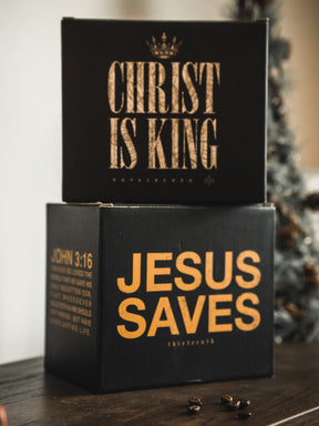 Two black boxes from 13th are stacked on a table. The top box features "CHRIST IS KING" in gold letters, while the bottom highlights the Jesus Saves Mug design with "JESUS SAVES" and John 3:16 in gold. A Christmas tree glows in the background.