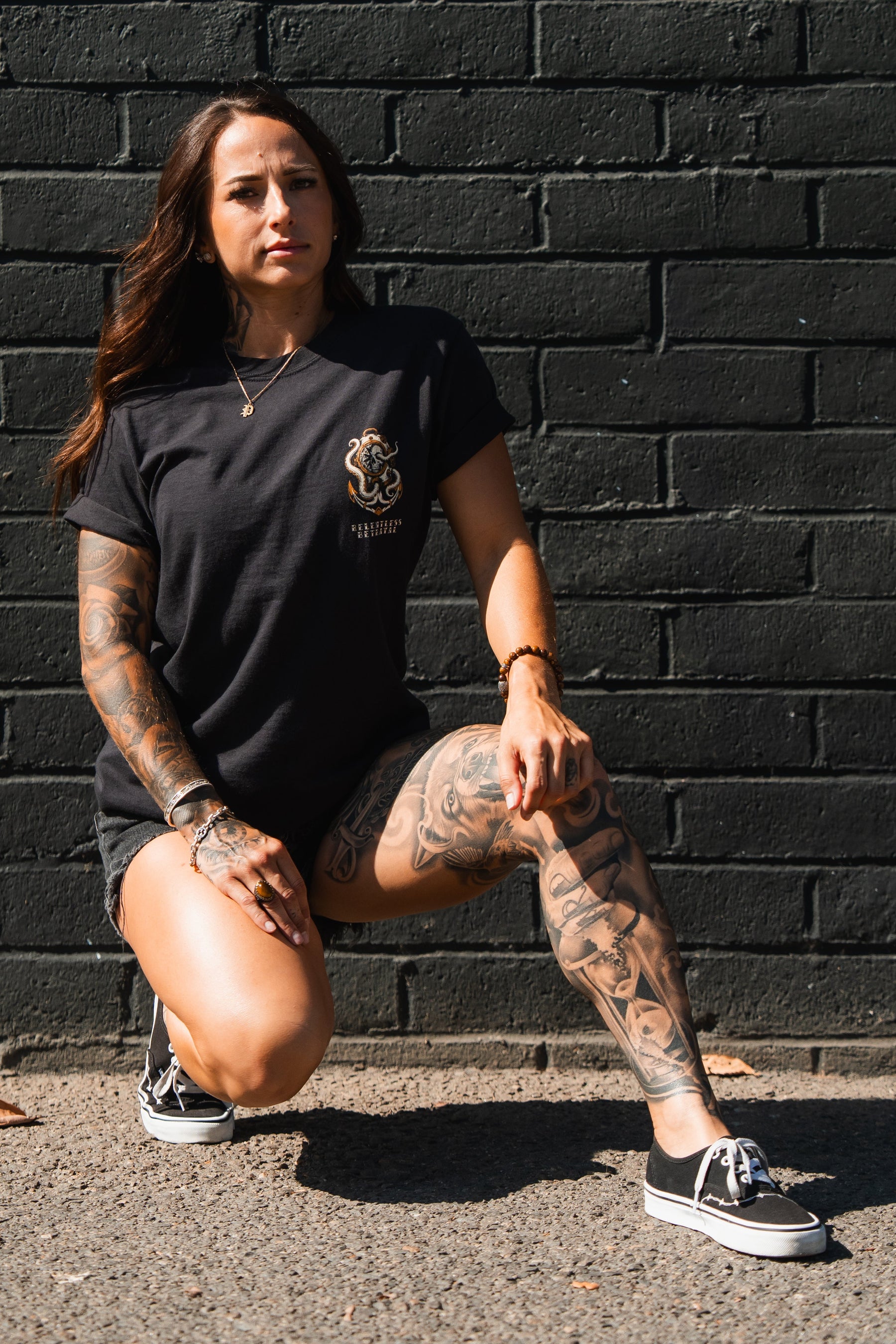 Tattooed woman kneels outdoors by a black brick wall, wearing the No Safe Harbor Premium T-Shirt from Relentless Betrayal, denim shorts, and black sneakers.