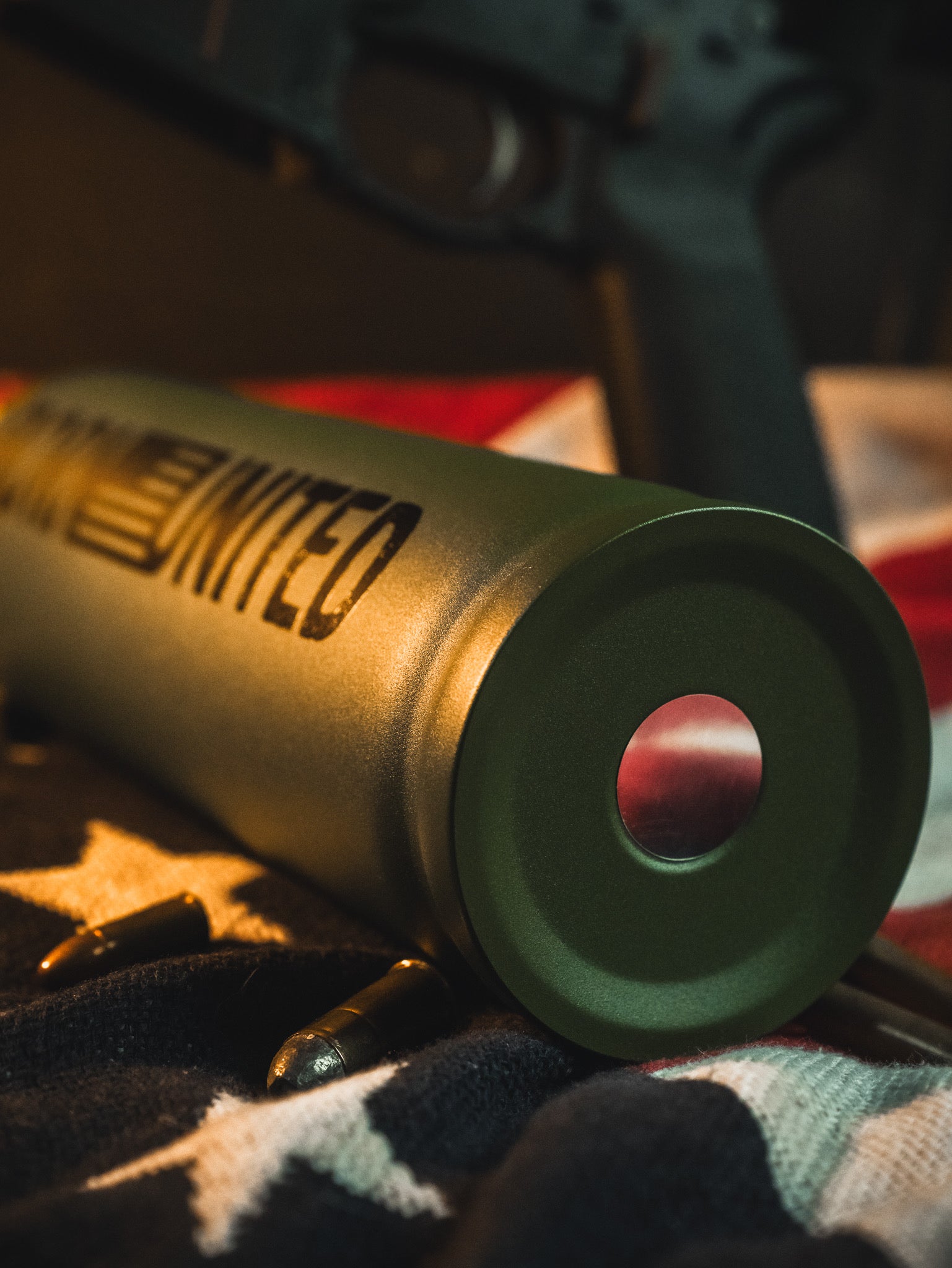 A close-up of a 32oz Born United Private Label Bullet Thermos with “United” and a US flag design, lying on an American flag with bullets nearby and part of a firearm visible in the background.