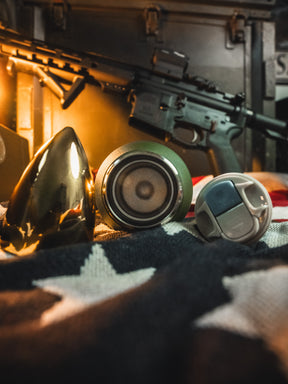 A shiny gold cone, a 32oz Born United Private Label Bullet Thermos, and a gray cap sit on a star-patterned cloth, with warm lighting and a blurred rifle in the background.